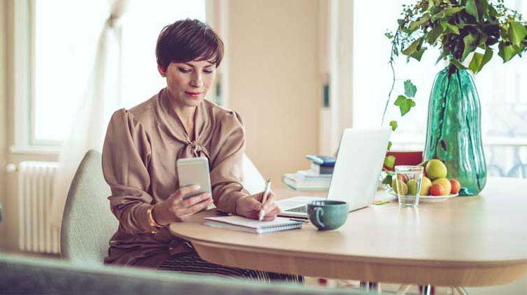 Woman sitting at a table, writing on paper while using her phone and laptop to research money market accounts.